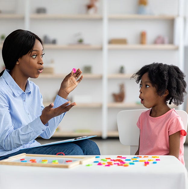 A woman explains something with hand gestures to a young child with magnet letters on a table between them.